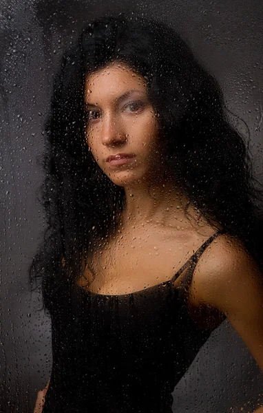 Hermosa chica detrás de vidrio húmedo sobre un fondo oscuro . — Foto de Stock