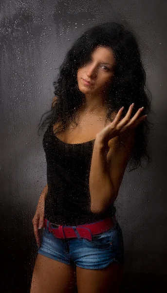 Hermosa chica detrás de vidrio húmedo sobre un fondo oscuro . — Foto de Stock