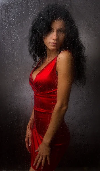 暗い背景にぬれたガラスの後ろに立っている、赤いドレスで美しい少女. — ストック写真