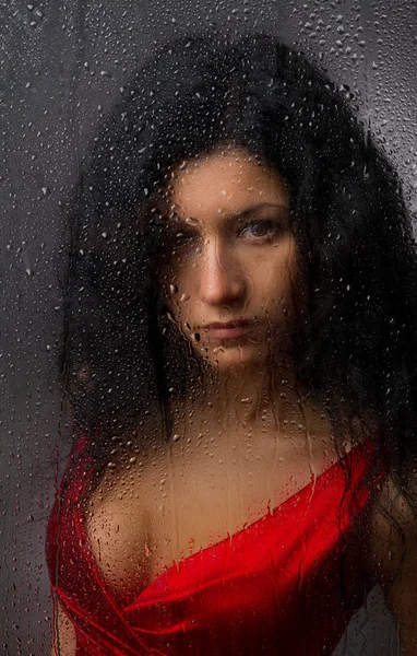 Belle fille dans une robe rouge, debout derrière un verre mouillé sur un fond sombre . — Photo