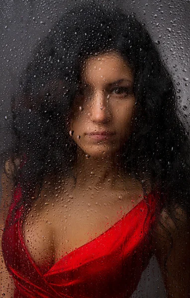 Beautiful girl in a red dress, standing behind a wet glass on a dark background. — Stock Photo, Image