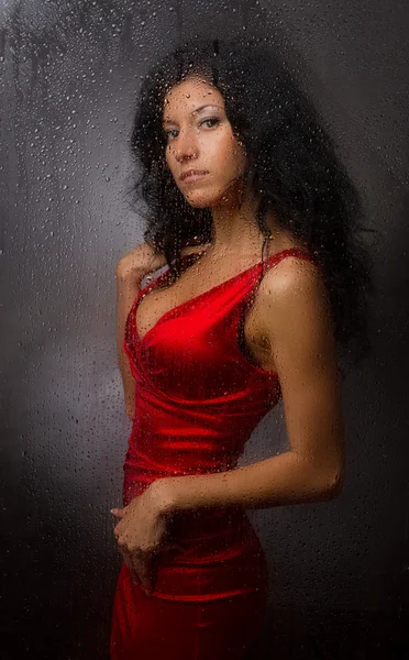 Vacker flicka i en röd klänning, står bakom en våt glas på en mörk bakgrund. — Stockfoto