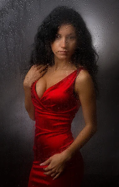 Beautiful girl in a red dress, standing behind a wet glass on a dark background. — Stock Photo, Image