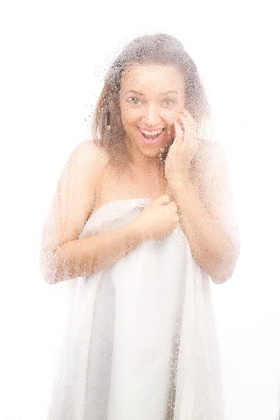 Schöne Mädchen in einem weißen Laken hinter einem nassen Glas. — Stockfoto