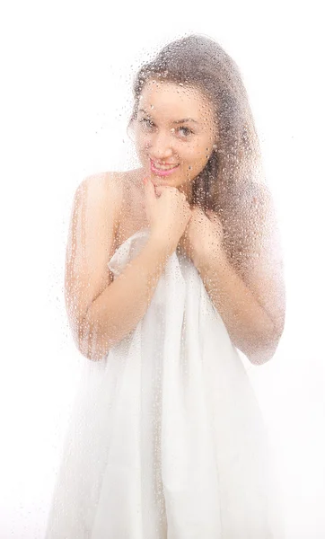 Hermosa chica en una sábana blanca detrás de un vaso mojado . — Foto de Stock