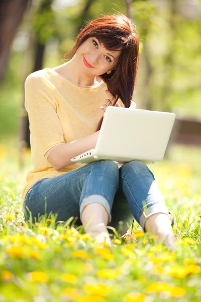 Roztomilá žena s bílým laptop v parku s pampelišky — Stock fotografie