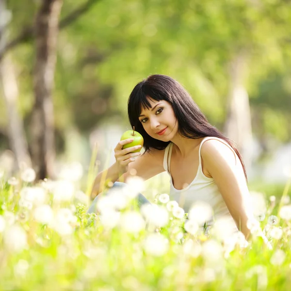 Cute woman eating the apple in the park with dandelions — Stock Photo, Image