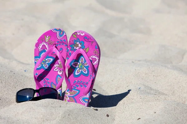 Ένα ζευγάρι ροζ χρώματος flip-flops στην άμμο με ένα μαύρο γυαλιά ηλίου — Φωτογραφία Αρχείου