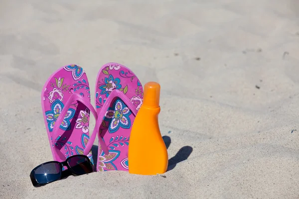 Ett par rosa färgade flip-flops i sanden med en svart solglasögon och sun mjölk — Stockfoto