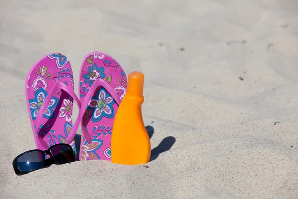 A pair pink colored Flip-Flops in the sand with a black sunglasses and sun milk — Stock Photo, Image
