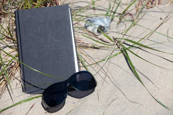 En mörka solglasögon ligger framme av en bok på en sanddyn — Stockfoto
