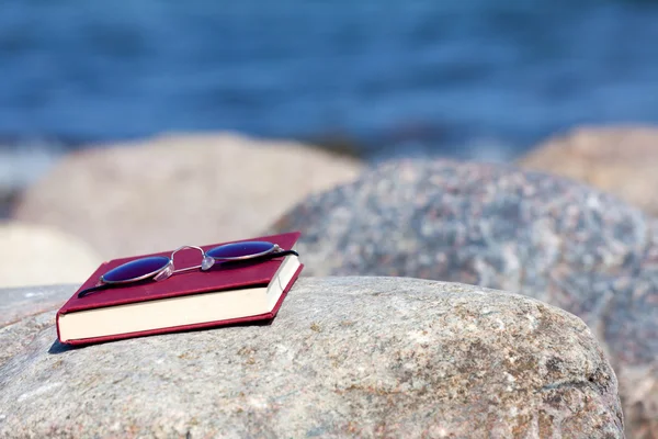 Libro cerrado con gafas de sol de color oscuro se encuentra en una roca en la playa —  Fotos de Stock