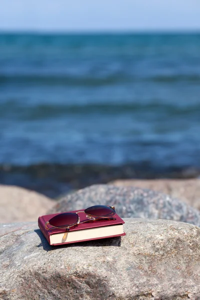 Fechado Livro com óculos escuros escuros está em uma rocha na praia — Fotografia de Stock