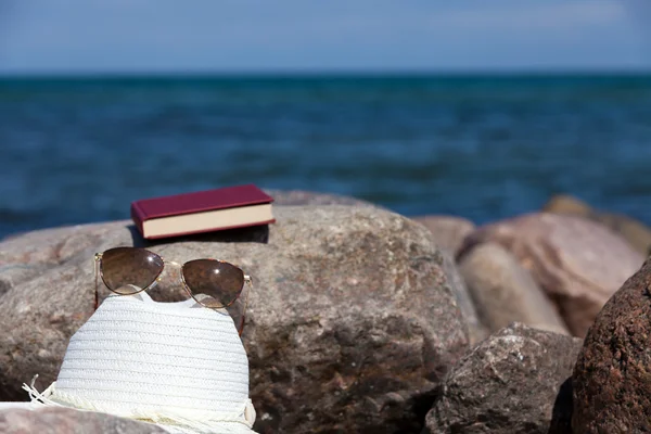 Zamkniętą księgą z okulary przeciwsłoneczne i kapelusz słomkowy leży na skale na plaży — Zdjęcie stockowe