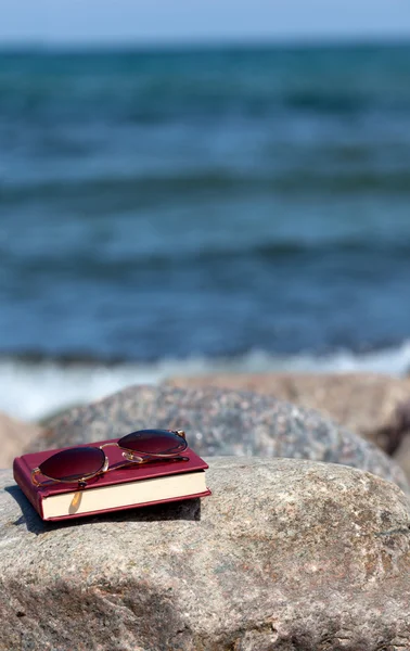 Закрита книга з темними тонованими сонцезахисними окулярами лежить на скелі на пляжі — стокове фото