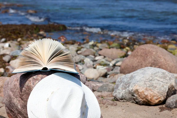 Um livro aberto mente e um chapéu de palha em uma rocha na praia — Fotografia de Stock