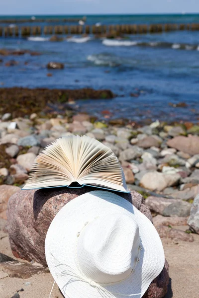 An opened book lies and a straw hat on a rock on the beach — Stock Photo, Image