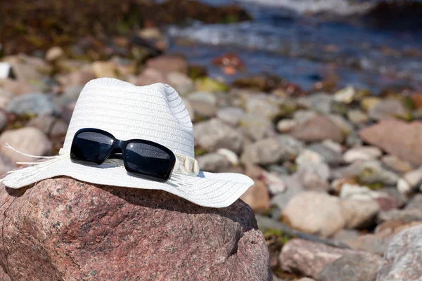 An white straw hat with sunglasses lies on a red big rock on the beach — Stock Photo, Image