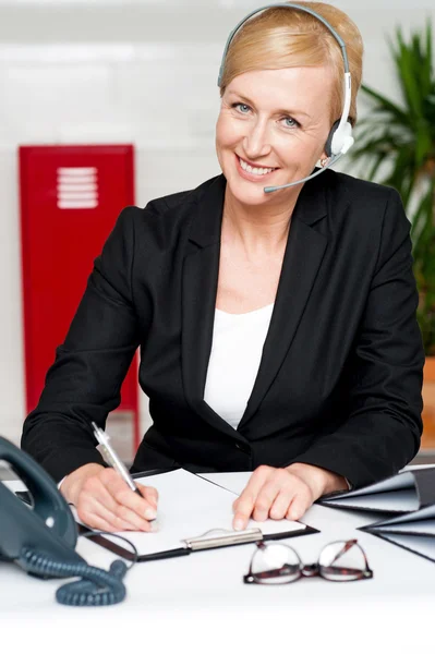 Mulher sorridente usando fone de ouvido escrevendo no bloco de notas — Fotografia de Stock