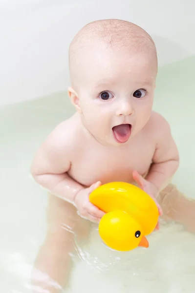 Pequeno bebê sorridente com pato amarelo sentado na banheira branca — Fotografia de Stock