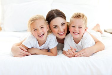 Laughing children playing with their mother lying on a bed clipart