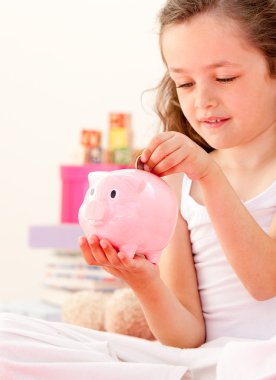 Cute little girl saving money in a piggy-bank sitting on her bed clipart