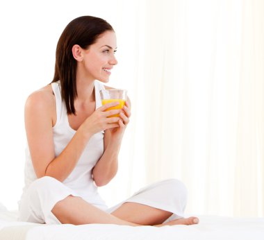 Cheerful woman drinking an orange juice sitting on her bed clipart