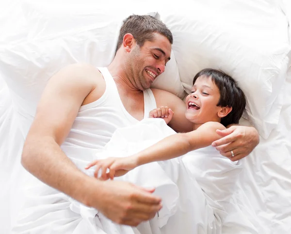 Ein lachender Junge spielt mit seinem Vater, der zu Hause auf einem Bett liegt — Stockfoto