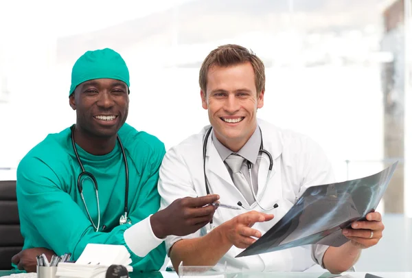 Νεαρός γιατρός και χειρούργος που είναι χαμογελώντας σε φωτογραφική μηχανή που κάθονται σε ένα γραφείο — Φωτογραφία Αρχείου