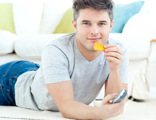 Relaxado jovem comendo batatas fritas segurando um controle remoto — Fotografia de Stock
