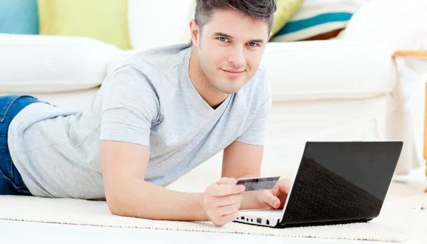 Smiling man lying on the floor with laptop holding a card — Stock Photo, Image