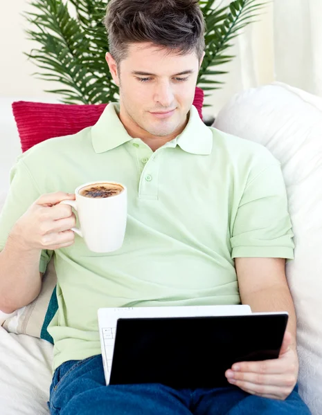 Χαμογελαστός νεαρός άνδρας, κρατώντας ένα φλιτζάνι καφέ που ψάχνετε σε ένα φορητό υπολογιστή — Φωτογραφία Αρχείου