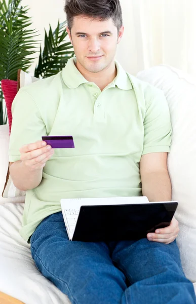 Χαρισματικός νεαρός με κάρτα και το φορητό υπολογιστή σε ένα καναπέ — Φωτογραφία Αρχείου