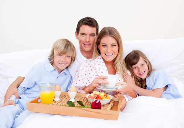 Leende familj äter frukost sitter på sängen — Stockfoto