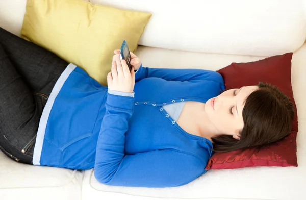 Mujer joven y relajada enviando un mensaje de texto tumbado en el sofá —  Fotos de Stock