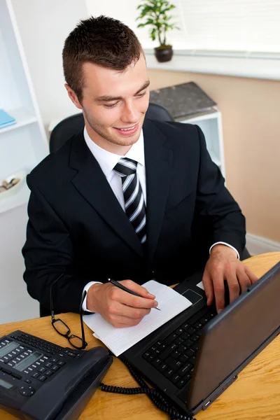 Joven hombre de negocios animado usando su computadora portátil escribiendo en un papel —  Fotos de Stock
