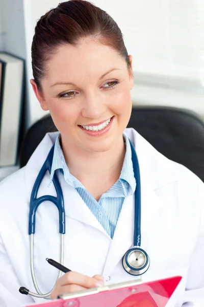 Glowing young doctor writing on a clipboard sitting in her offic — Stock Photo, Image