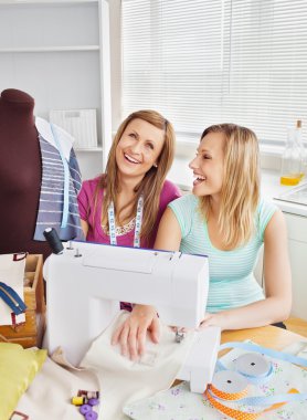 Laughing female friends sewing clothes together at home clipart