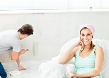 Smiling caucasian woman relaxing on a sofa while boyfriend paint clipart