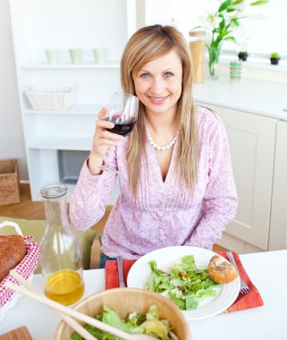 Bright young woman drinking wine and eating a salad in the kitch clipart