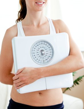 Close-up of a fit hispanic woman holding a scale in her living-r clipart