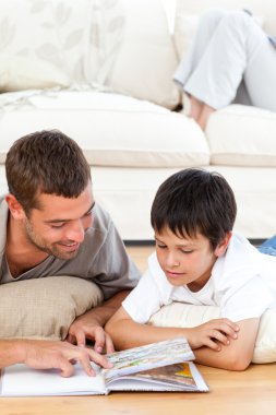 Attentive father reading a book with his son lying on the floor clipart