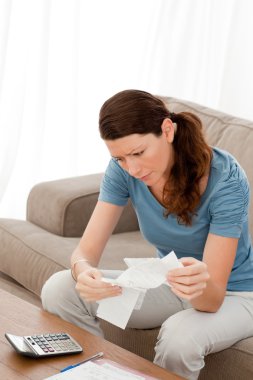 Worried woman looking at her bills sitting on her sofa clipart