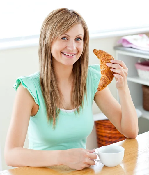 Mujer joven animada comiendo un croissant sosteniendo una taza en el kit —  Fotos de Stock