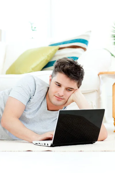Очаровательный молодой человек использует свой ноутбук, лежащий на полу — стоковое фото