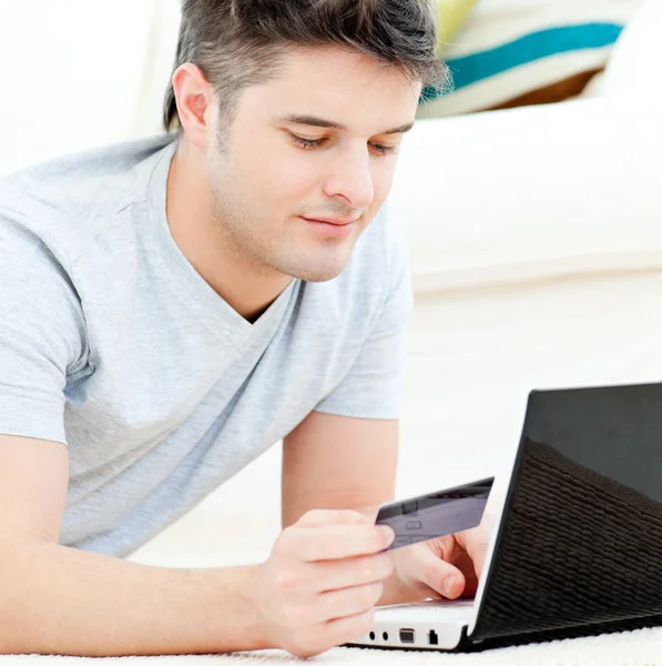 Positive young man holding a card using his laptop on the floor — Stock Photo, Image