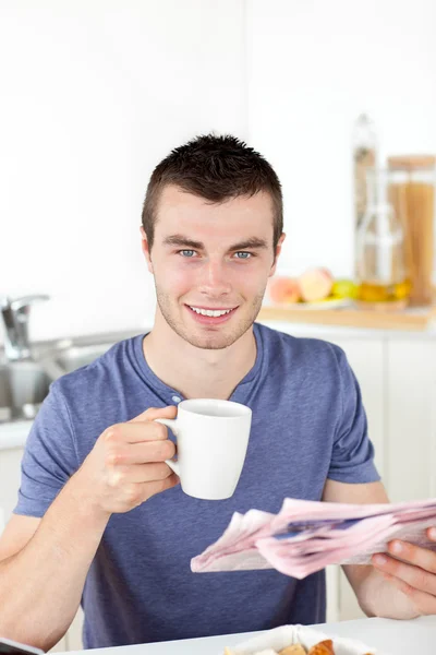 Joven positivo sosteniendo una taza y un periódico sonriendo al — Foto de Stock