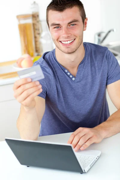 Charismatic young man holding a card using his laptop in the kit — Stock Photo, Image
