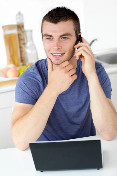 Χαμογελαστός άνθρωπος που μιλάει στο τηλέφωνο χρησιμοποιώντας το φορητό υπολογιστή στην κουζίνα — Φωτογραφία Αρχείου