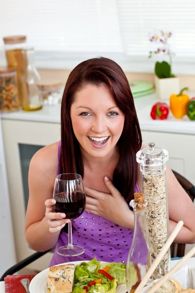 Mulher alegre comendo sua refeição segurando um copo de vinho em casa — Fotografia de Stock